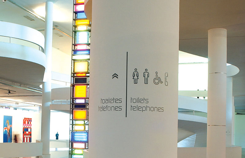 Sinalização de serviços da 26ª Bienal de São Paulo no Pavilhão Ciccillo Matarazzo, com pictogramas e indicação bilíngue de toaletes e telefones integrada à arquitetura.