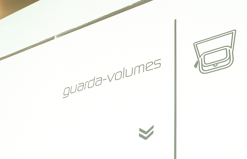 Sinalização de guarda-volumes da 26ª Bienal de São Paulo, com indicação tipográfica, seta direcional e pictograma aplicados ao sistema de wayfinding da exposição.