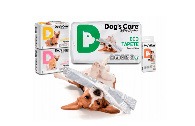 Cachorro deitado segurando tapete com logos Dog's Care, embalagens de fraldas nas cores amarela e rosa, e caixa de eco tapete ao fundo