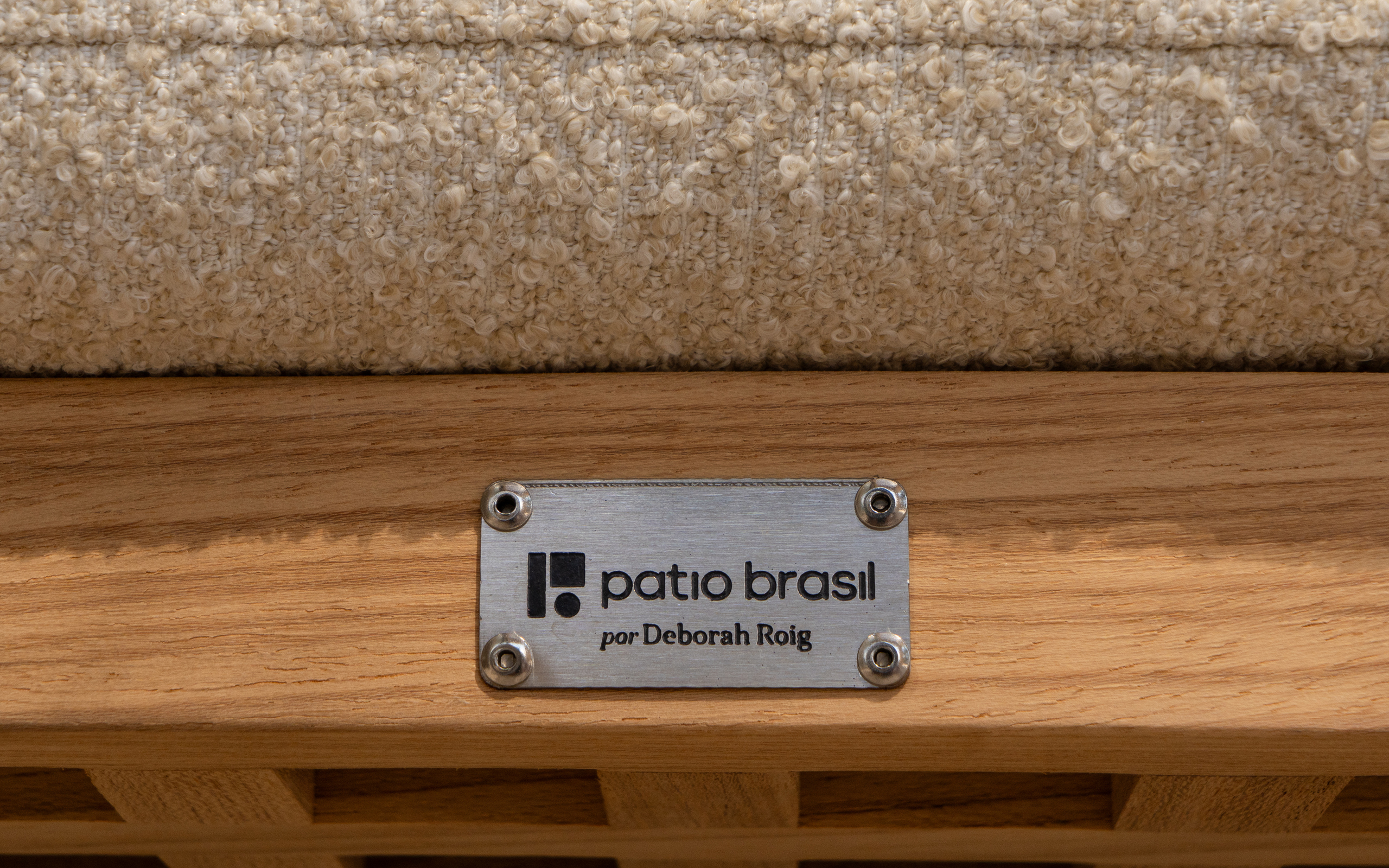 Patio Brasil metal plaque attached directly to the wooden structure of an outdoor furniture piece, featuring the symbol, wordmark and “por Deborah Roig” designer credit. The application demonstrates the visual identity physically integrated into the product and its designer attribution.